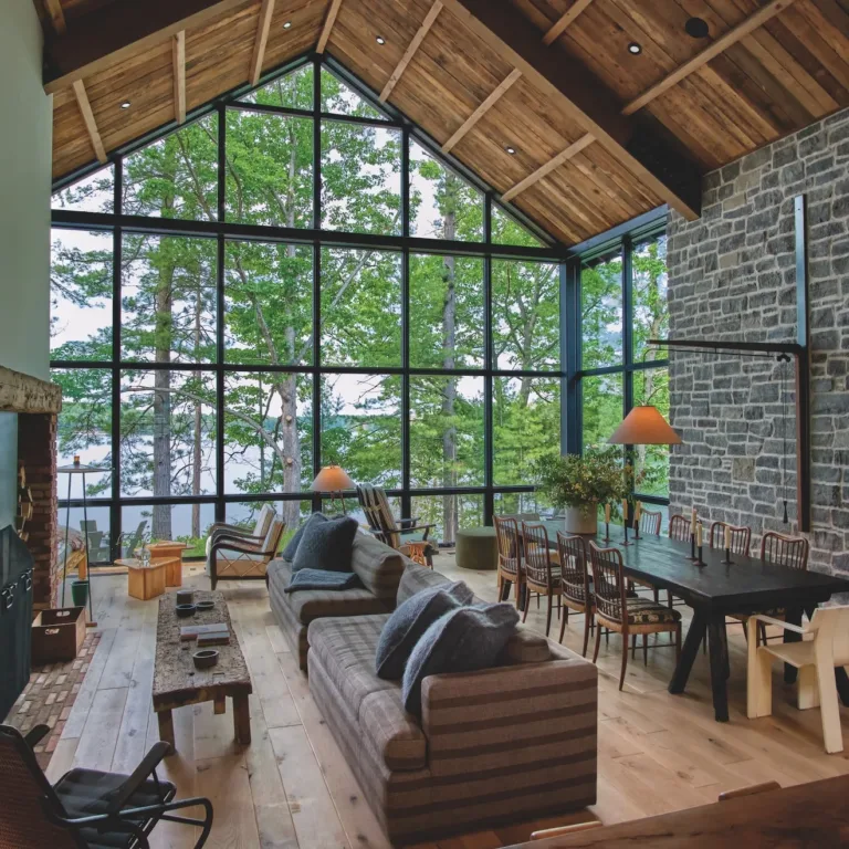 Spacious living and dining area with large floor-to-ceiling windows showing outdoor trees and lake, featuring a stone wall, wooden ceiling, brick fireplace, striped sofas, wooden dining table with chairs, and pendant lamps.