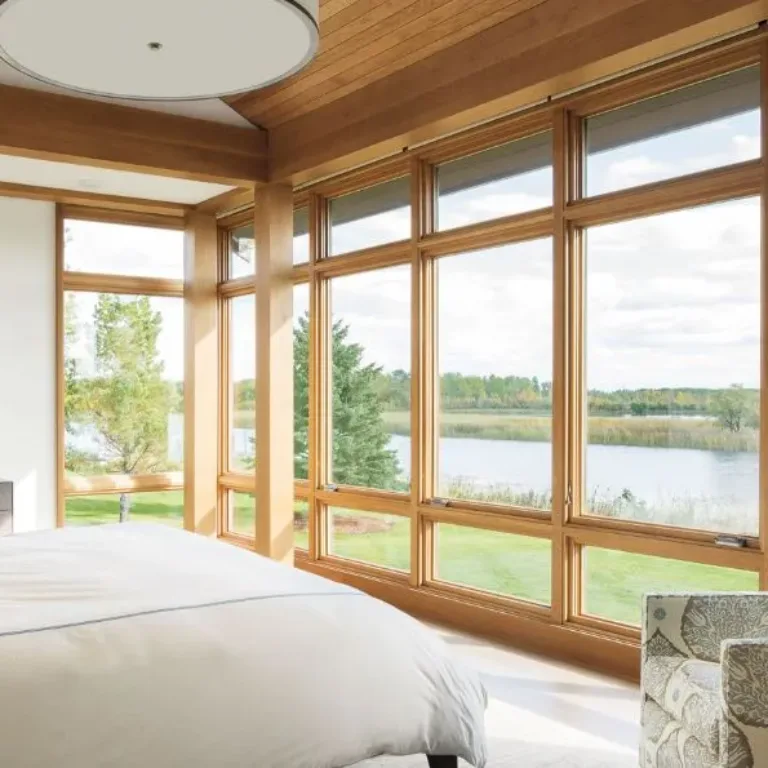 Bright bedroom with large floor-to-ceiling wooden-framed windows overlooking a lake and greenery, featuring a white bed and patterned armchair.