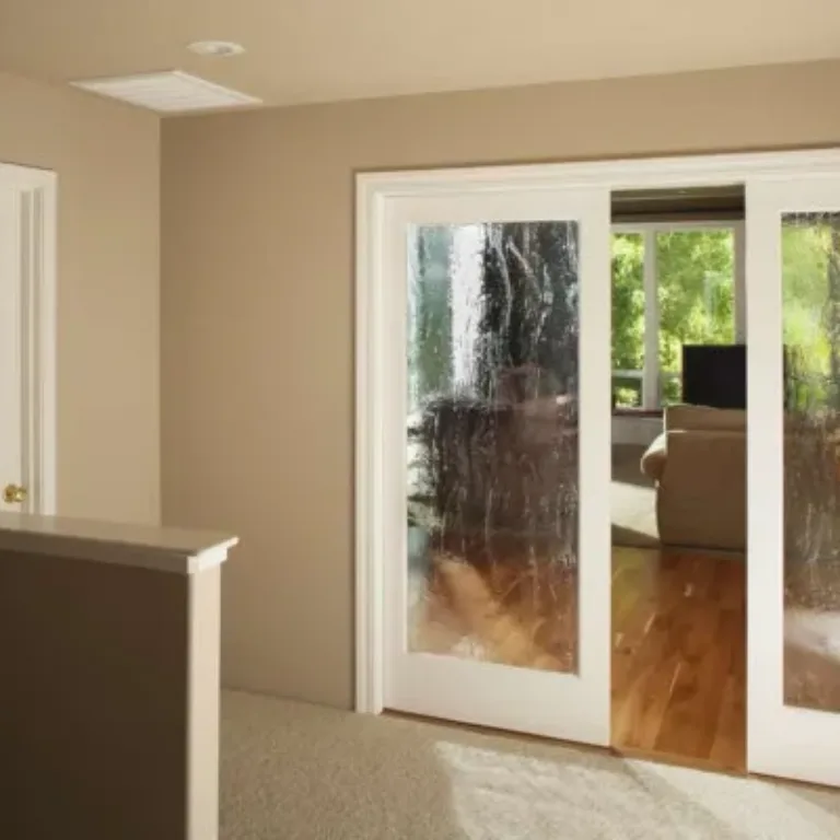 Interior view of a beige room with carpeted floor and a half wall leading to a space with white-framed sliding doors featuring textured glass panels, partially open to reveal a living area with wooden flooring, a beige couch, and large windows with green trees outside.