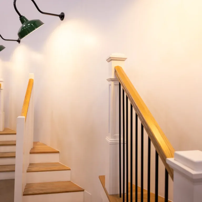 Three sets of indoor wooden stairs with white railings and light wood handrails, illuminated by green wall-mounted lamps against a white wall.