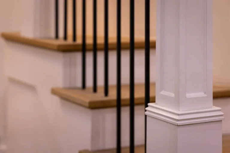 Close-up of a white wooden bannister post with decorative molding next to a staircase with wooden steps and black vertical metal balusters.
