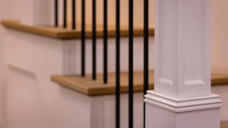 Close-up of a white wooden bannister post with decorative molding next to a staircase with wooden steps and black vertical metal balusters.