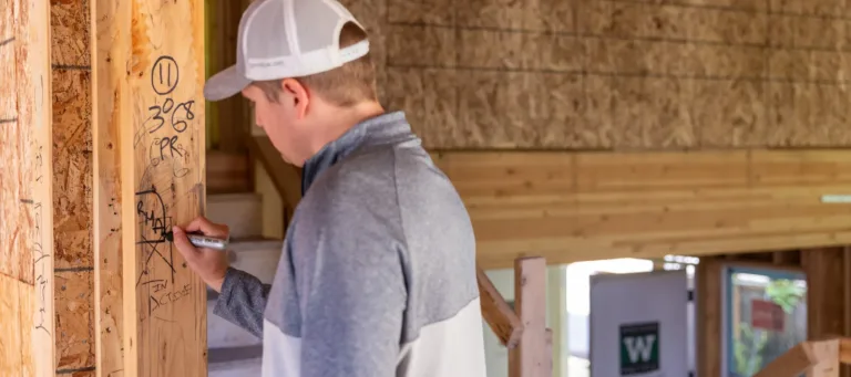 Person wearing a gray and white jacket and light gray baseball cap writing on a wooden beam inside a building under construction.