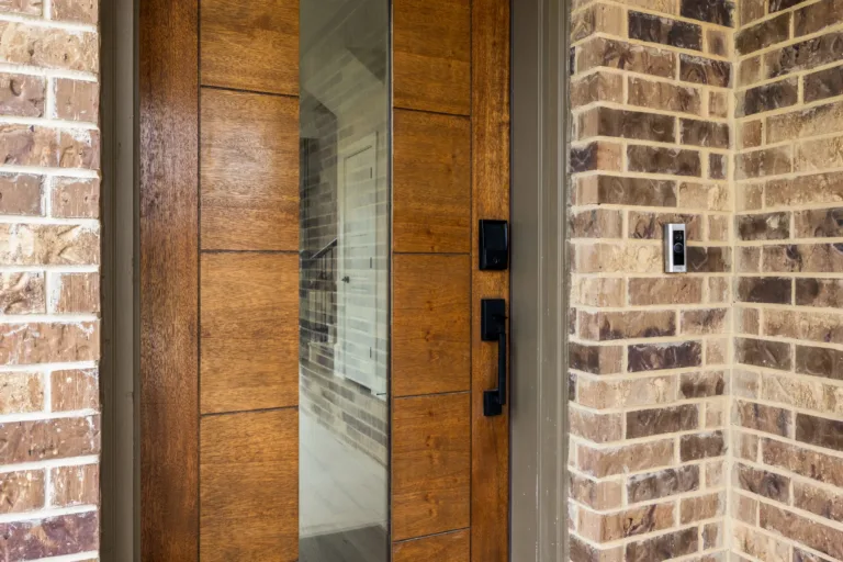 Wooden front door with vertical glass panel and black handle on a brick house exterior featuring a ring doorbell on the right brick wall.