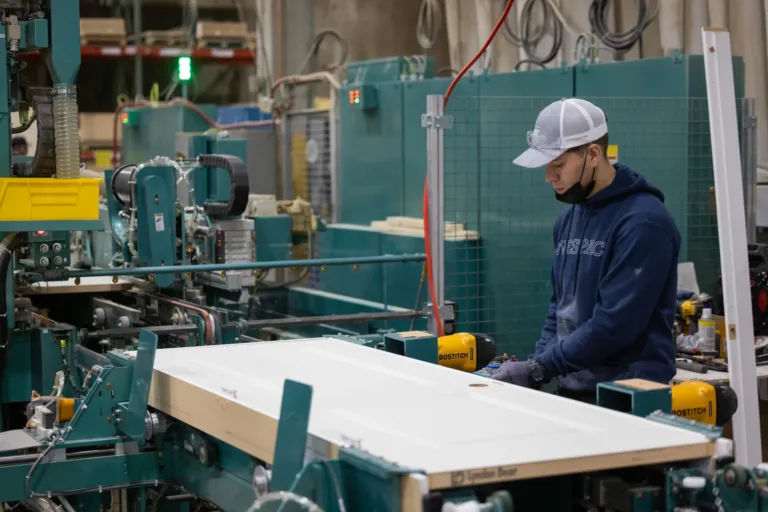 Worker wearing a gray cap, navy hoodie, gloves, and safety goggles operating machinery to assemble a large white door in an industrial workshop.