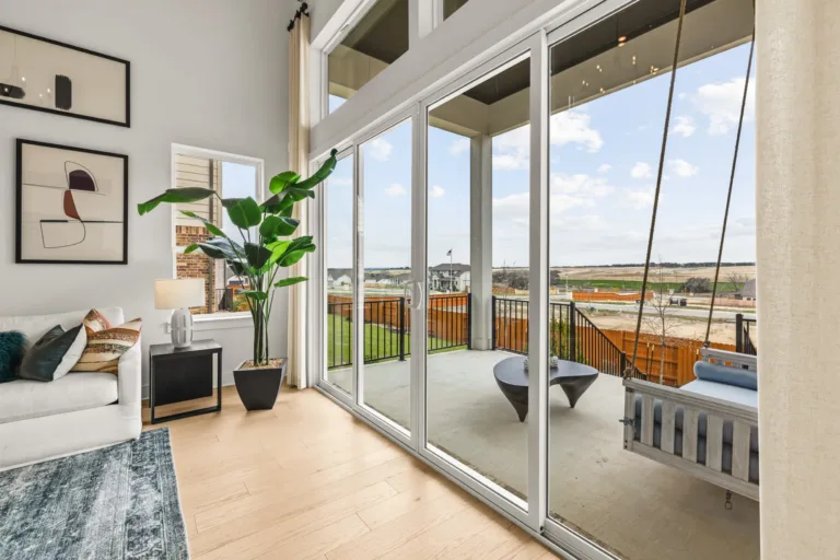 Modern living room with white sofa adorned with colorful pillows, a large indoor plant in a black pot, abstract wall art, and large glass sliding doors leading to an outdoor patio featuring a swing bench and a black coffee table, overlooking a suburban neighborhood and open landscape.