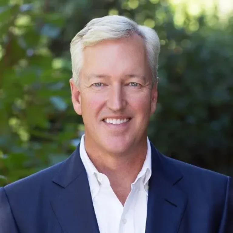 Portrait of a middle-aged man with short white hair wearing a dark blazer and white shirt, smiling against a blurred green outdoor background