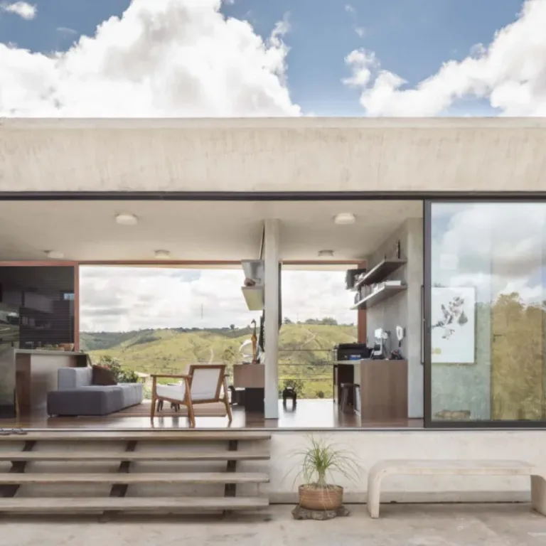 Modern open-plan living space with large glass walls, a gray sofa, wooden chairs, and a view of green hills, with concrete stairs and outdoor plants in front.