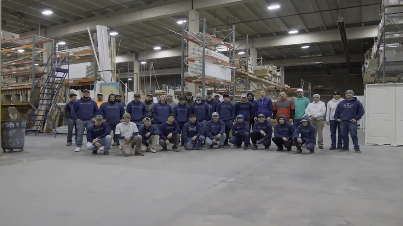 Group photo of approximately 30 workers in a warehouse with shelves holding construction materials, all wearing casual work attire including hoodies, jackets, and caps, standing and crouching on a concrete floor under industrial lighting.