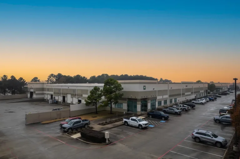 Large industrial warehouse building with multiple loading docks, parking lot filled with cars and trucks, two trees near the building corner, under a clear sky at sunset.