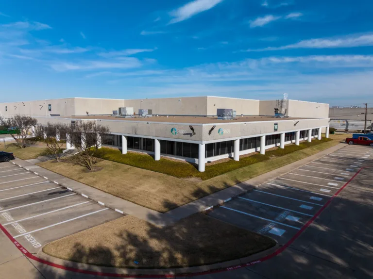 Exterior view of a large beige commercial building with white columns and a flat roof, surrounded by dry grass landscaping and mostly empty parking spaces under a clear blue sky.