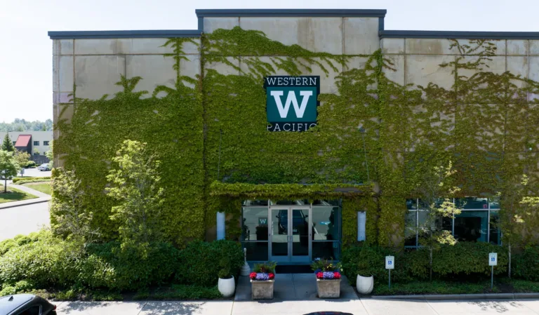 Entrance of a concrete building covered with green ivy, displaying a sign for Western Pacific above double glass doors, with potted plants and parking spaces visible in front.