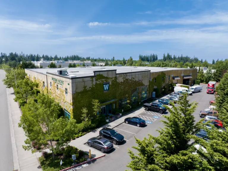 A large, rectangular commercial building covered partly with green vines, named Western Pacific, with a parking lot featuring various cars including black SUVs and a red truck, surrounded by trees and clear blue sky above