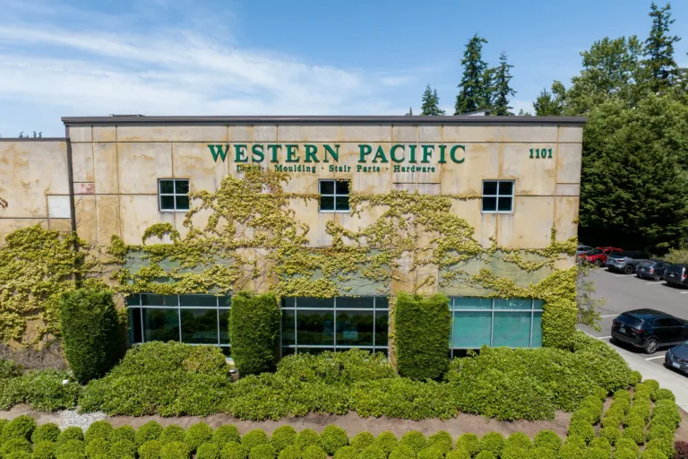 Exterior of a beige commercial building with green ivy growing on the facade, displaying the sign 'Western Pacific Moulding Stair Parts Hardware' and the number 1101, surrounded by green landscaping and parked cars under a blue sky