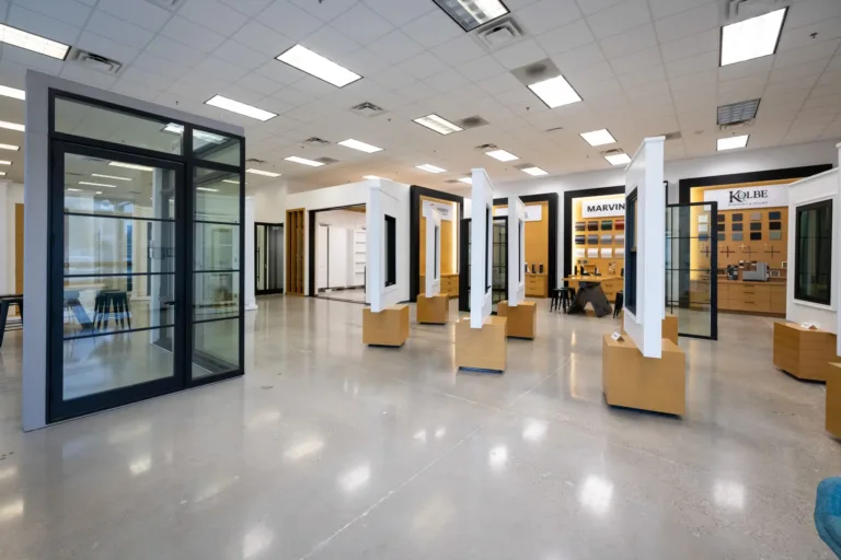 Bright and spacious showroom displaying various window and door models mounted on wooden stands with brand signs like Marvin and Kolbe in the background, featuring polished concrete floors and ceiling lights.