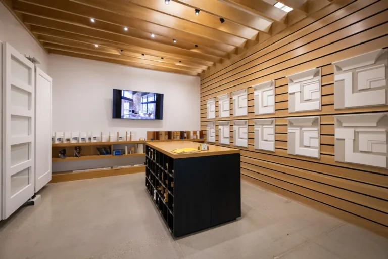 Interior of a modern showroom with wooden slat walls displaying white architectural molding samples, a central black storage island with wood top, and a large flat-screen TV mounted on a white wall under a wood-beamed ceiling with recessed lighting.