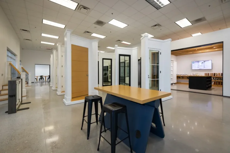 A modern showroom featuring a central table with a wooden top and dark blue base surrounded by black stools, with multiple white door frames displaying different door styles in the background and a well-lit adjacent room with a TV and wooden ceiling beams.