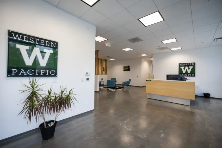 Modern office reception area with polished concrete floors, white walls displaying Western Pacific signs, a wooden reception desk with computer monitors, a potted plant near the entrance, and a seating area with blue chairs and a table in the background.