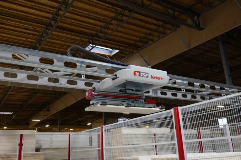 Industrial robotic arm labeled CSF barbaric mounted on a ceiling rail system inside a warehouse, positioned above fenced storage areas with a wooden ceiling in the background