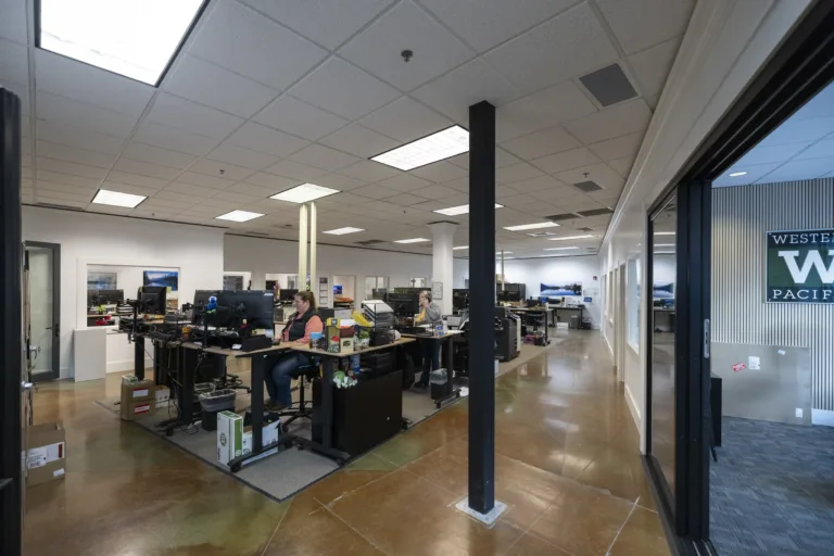 Open office space with multiple desks and computer monitors, two people working, polished concrete floor, white walls, ceiling lights, and a glass-walled room on the right with a green sign that reads 'Western Pacific'.
