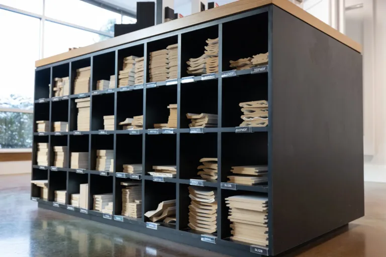 Black cubby storage unit with cubicles holding neatly stacked wooden molding samples, each compartment labeled with codes, in a bright room with large windows and a polished floor.