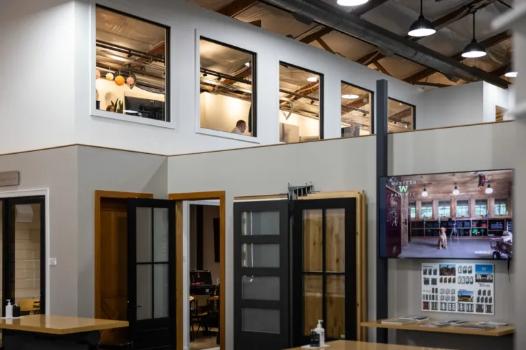 Interior view of an office space with white walls, black-framed windows showing people working inside, open wooden doors, and a mounted flat-screen TV displaying an image of a rustic workshop with a dog.