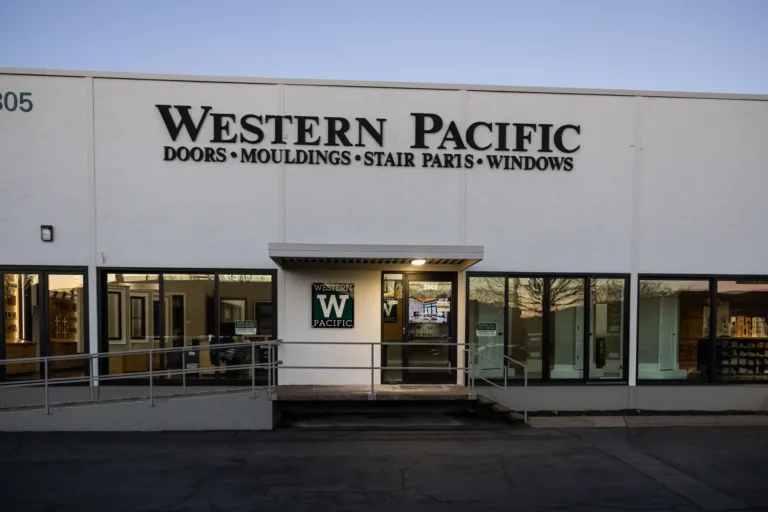 Exterior view of a white commercial building with large black lettering reading 'Western Pacific Doors Mouldings Stair Parts Windows' above the entrance and several large windows on either side of the door.