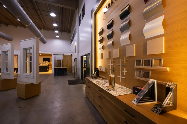 Interior of a showroom with various architectural display samples mounted on a wooden wall and countertop, including curved metal sheets, wooden panels, architectural frames, and cross sections of window or door frames, with high ceilings, industrial lighting, and open floor space featuring window display models on wooden platforms.