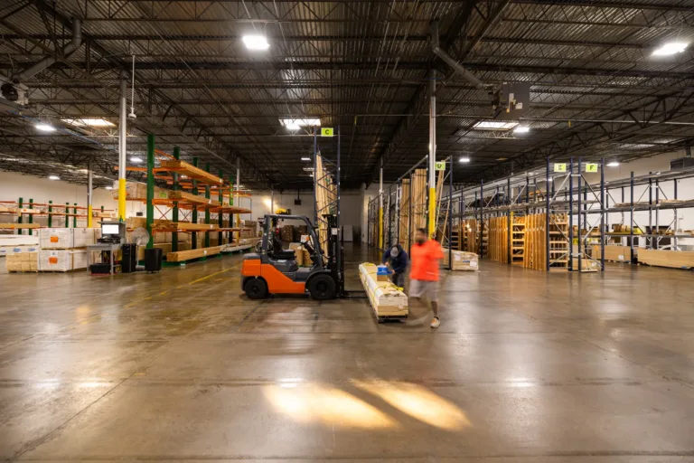 Large warehouse interior with high ceilings, containing shelves stocked with wooden planks and materials, an orange forklift carrying a load of wood, and two workers in motion near the forklift, one wearing a bright orange shirt.