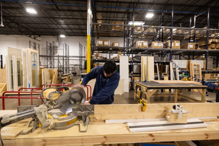 Person wearing a navy sweatshirt using a miter saw to cut wood in a large workshop filled with shelves, wooden doors, and various woodworking materials and tools.