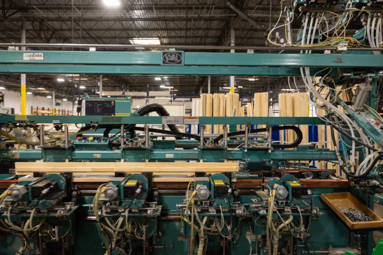 A woodworking machine in a factory holding multiple wooden planks, surrounded by various tubes, wires, and mechanical components with stacks of wooden boards in the background.