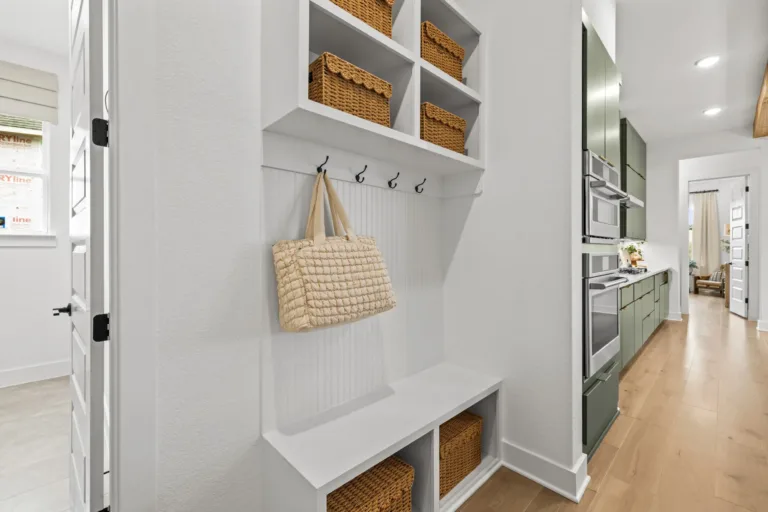 White built-in storage unit with cubbies holding wicker baskets, black hooks with a beige quilted bag hanging, adjacent to a kitchen with green cabinetry and stainless steel appliances, light wooden flooring throughout.