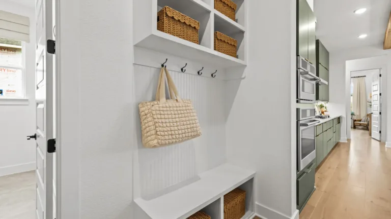 White built-in storage unit with cubbies holding wicker baskets, black hooks with a beige quilted bag hanging, adjacent to a kitchen with green cabinetry and stainless steel appliances, light wooden flooring throughout.