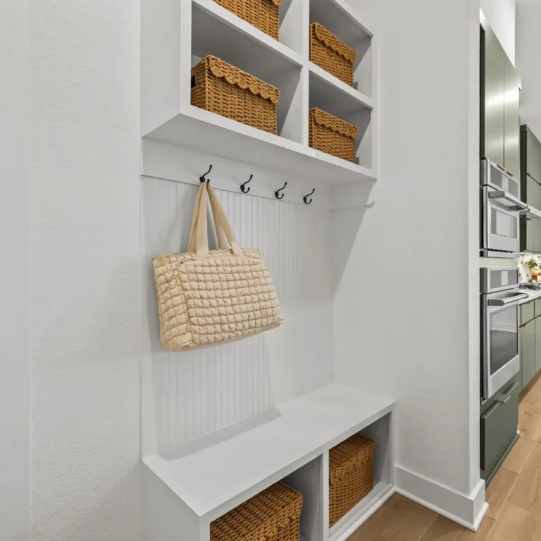 White built-in storage unit with cubbies holding wicker baskets, black hooks with a beige quilted bag hanging, adjacent to a kitchen with green cabinetry and stainless steel appliances, light wooden flooring throughout.