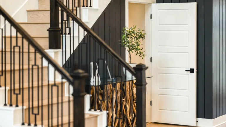 Indoor staircase with light wooden steps and black metal railing next to a white door set in a dark paneled wall, with a small potted plant partially visible through the open door.