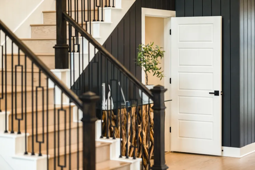 Indoor staircase with light wooden steps and black metal railing next to a white door set in a dark paneled wall, with a small potted plant partially visible through the open door.