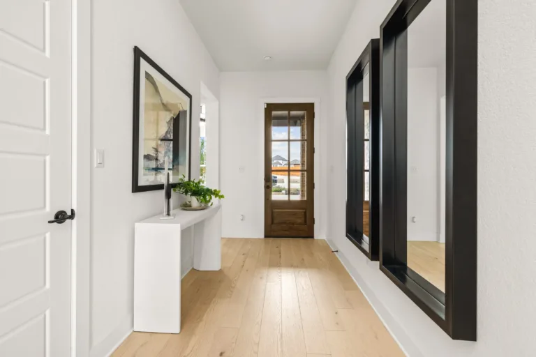 Minimalist hallway with light wood flooring, white walls, a wooden door with glass panels at the end, a white console table with a plant and decorative object, a framed abstract artwork on the left wall, and two large black-framed mirrors on the right wall.