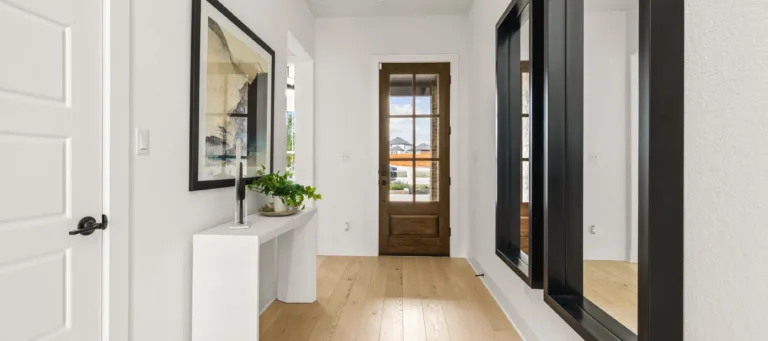 Minimalist hallway with light wood flooring, white walls, a wooden door with glass panels at the end, a white console table with a plant and decorative object, a framed abstract artwork on the left wall, and two large black-framed mirrors on the right wall.