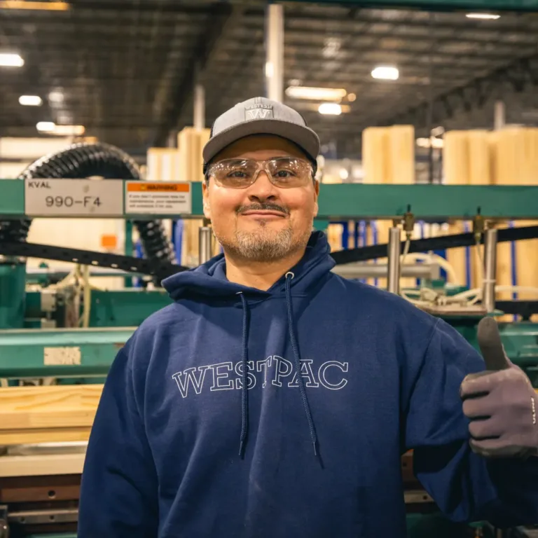 Smiling man wearing safety glasses, a gray cap, and a navy hoodie with "WESTPAC" printed on it, giving a thumbs-up inside an industrial workshop with machinery and wooden materials in the background.