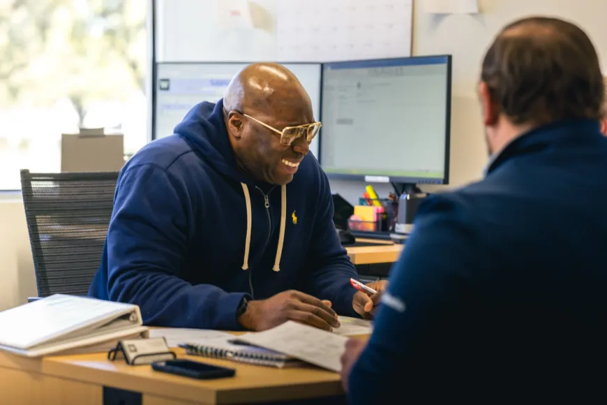 Two men sitting at a desk in an office, one man wearing a navy hoodie and glasses is smiling and holding a pen, the other man is seen from behind wearing a dark blue shirt, with papers, a notebook, and a smartphone on the desk and computer monitors in the background.