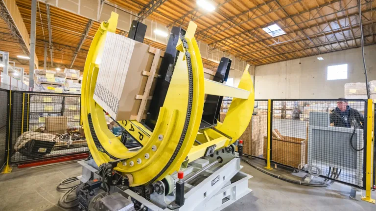 Industrial robotic arm with yellow curved clamps holding a wooden panel inside a warehouse with a worker operating a control panel behind a safety fence.