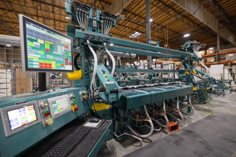 Industrial machine with multiple screens, control panels, and tubes in a large warehouse with wooden ceiling and shelves in the background.