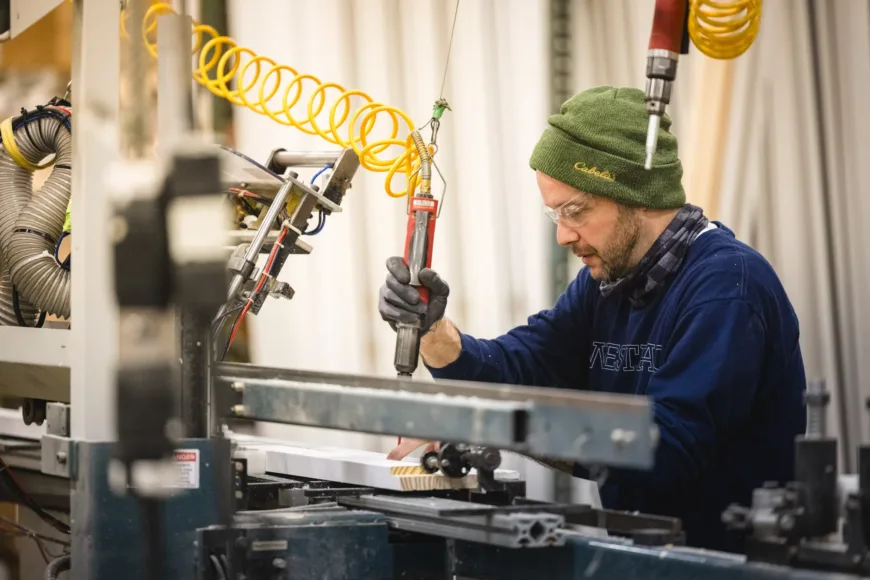 Man wearing a green beanie and safety glasses operating woodworking machinery in a workshop.