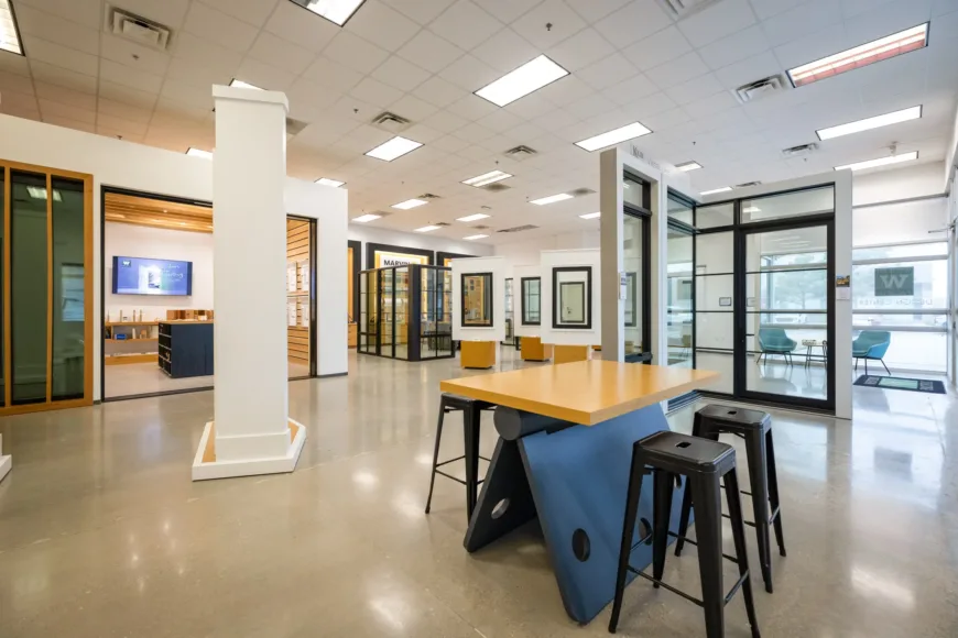 Modern interior showroom with polished concrete floor, high ceiling with fluorescent lights, glass window and door displays, a blue A-frame table with a wooden top, and black stools around it.