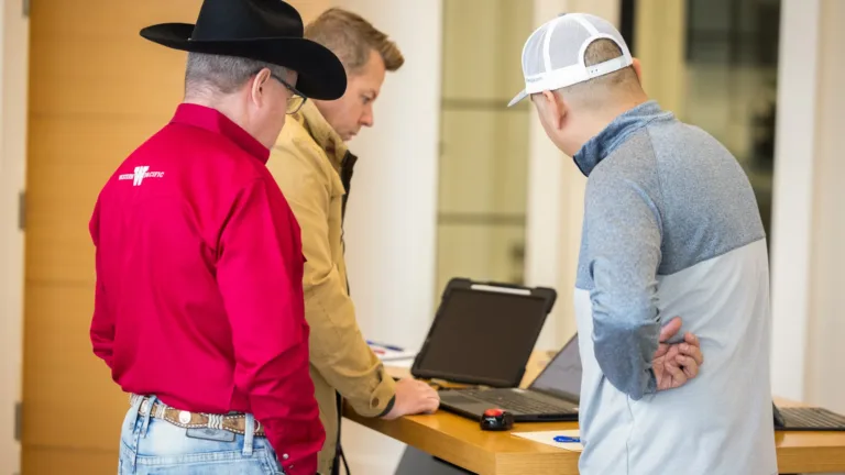 Three men standing around a wooden table looking at laptops, one wearing a red shirt and black cowboy hat, another in a tan jacket, and the third in a gray and white jacket with a white mesh cap.