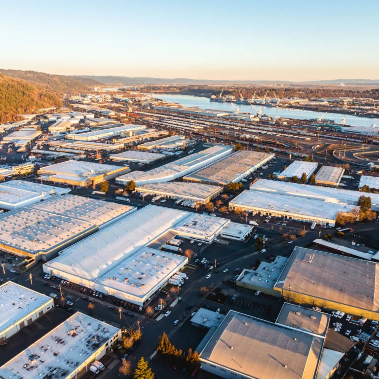 Aerial view of an industrial area with numerous warehouses featuring white and gray roofs, adjacent to a river with cranes and shipping docks, and surrounded by forested hills under a clear sky at sunset.