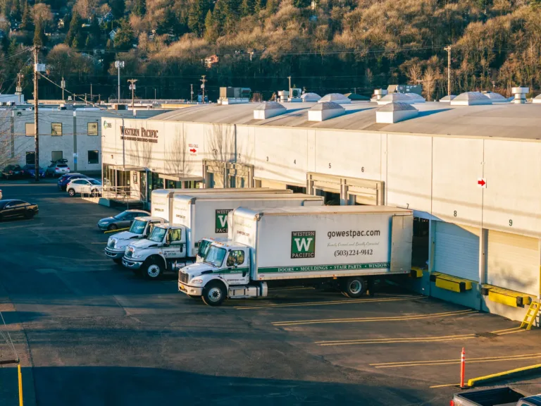 Three white Western Pacific delivery trucks parked in loading bays outside a large warehouse building with numbered bays, surrounded by parked cars and trees on a hillside in the background.