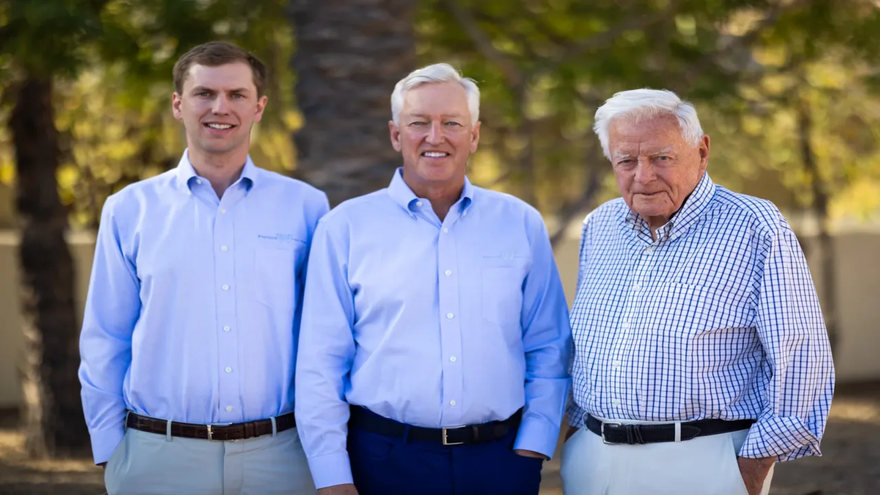 Three men standing outdoors in front of blurred trees, all wearing button-up shirts; the younger two men wear light blue shirts and light-colored pants, while the older man wears a white and blue checkered shirt with light pants.