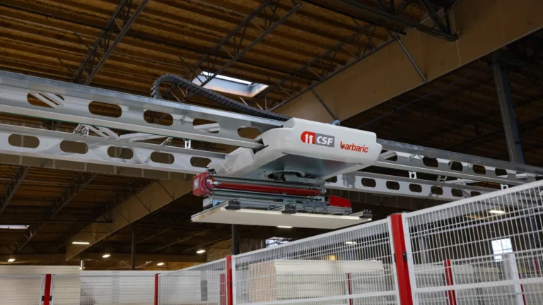 Automated overhead robotic system labeled 'CSF barbaric' inside an industrial warehouse, used for material handling or manufacturing processes, with metal framework and fenced enclosures below.