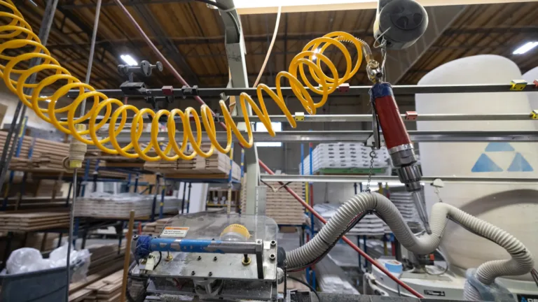 Industrial woodworking machinery with a red pneumatic tool attached to a yellow coiled hose in a workshop filled with wooden boards and storage racks.